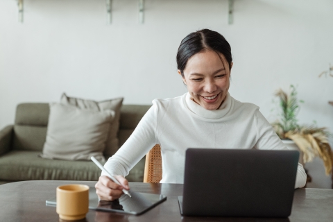 Woman smiling at her laptop while taking notes on a tablet