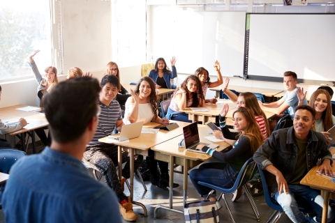 High school students looking at teacher raising their hands in a classroom